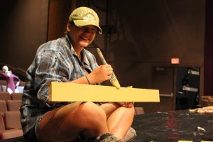 Photo of a teenage intern seated on the stage floor painting a wooden board and smiling toward the camera.