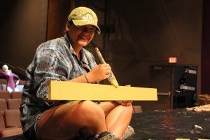 Photo by Cindy Dauer Photo of a teenage intern seated on the stage floor painting a wooden board and smiling toward the camera.