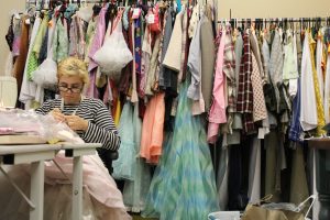 Photo by Cindy Dauer Photo of an intern sewing at a table with a large 2-tiered rack of costumes behind them.