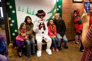 Photo from the event of a family of two daughters, mom and dad, all seated with Frosty the Snowman in the theatre lobby in front of a backdrop of a snow-covered hill.