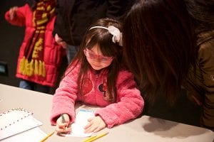 Photo from the event of a little girl with dark hair, pink glasses, and a puffy pink jacket thoughtfully writing something down on a piece of paper.