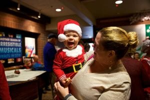 Photo from the event of a woman holding a toddler that is wearing a santa costume complete with little hat. The toddler smiles with their mouth wide open, having a good laugh.