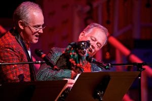 Photo from the event of two men on stage singing. The man on the right is holding a small black dog that is smiling and wearing a large green and red color with jingle bells on it.
