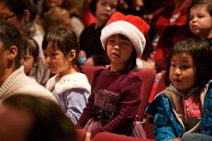 Photo from the event of three children seated in the theatre, the one in the middle wearing a Santa hat. All three watch the stage with wonder.