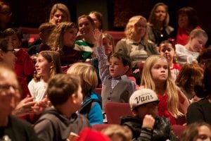 Photo from the event of the audience seated in the theatre. In the center of the photo is a small boy dressed in a plaid shirt, vest, and bowtie, raising his right hand high over his head.
