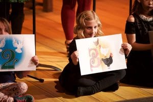 Photo from the event of a small girl on seated on the stage floor holding a sign with an elegantly drawn number three that also has three chickens surrounding the number.