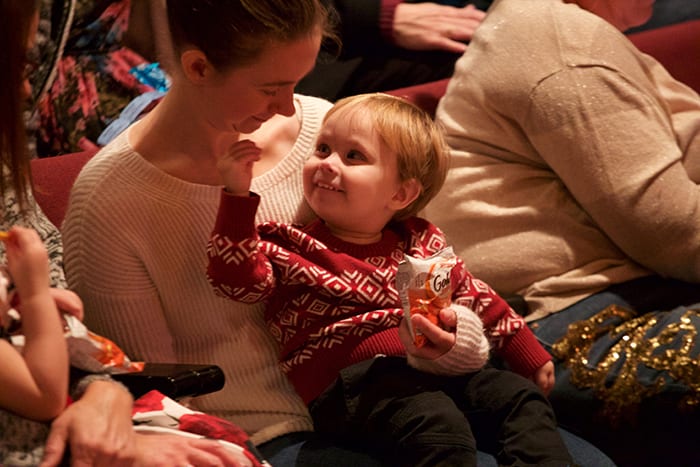 Photo from the event of a woman seated in the theatre holding a toddler in her lap. The toddler is paqrtially turned around toward the woman and smiling with a big, toothy grin.