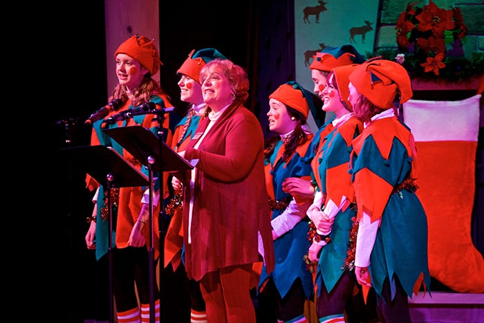 Photo from the event of a woman on stage with a music stand in front of her. Behind her is a small chorus of "Elves" dressed in green and red costumes.