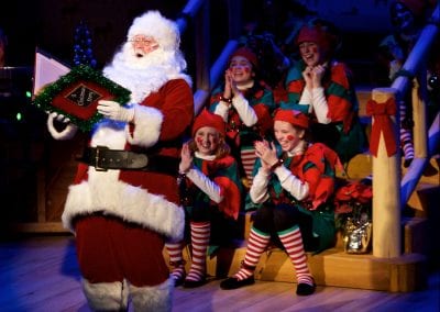 Photo of Santa on stage reading with a group of Santa's helpers listening behind him.