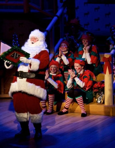 Photo of Santa on stage reading with a group of Santa's helper's listening behind him.
