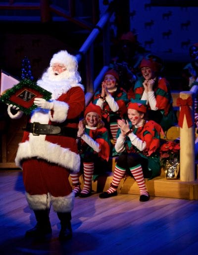 Photo of Santa on stage reading with a group of Santa's helper's listening behind him.