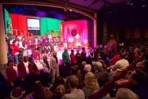 Photo from the event of man people singing, both onstage and down in front of the first row. They are all dressed festively in front of the brightly lit set.