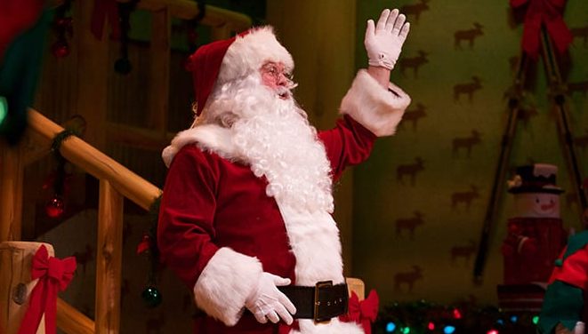 Photo of Santa Claus waving to the audience from stage. He wears his traditional red and white suit with white gloves and a big black belt.