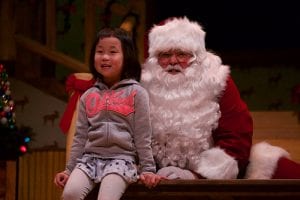 Photo from the event of Santa and a little girl, side by side. She is seated on a wooden banister railing in front of Santa Claus.