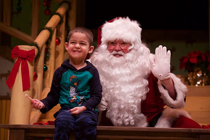 Photo from the event of a little boy seated in front of Santa Claus, smiling big. He is seated on a wooden banister rail in front of Santa who is smiling and waiving.