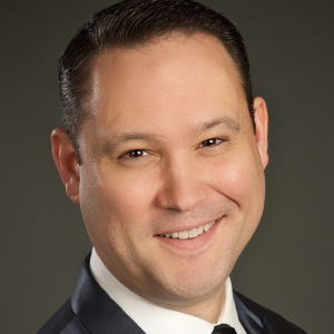 Smiling man with brown eyes and dark hair wearing a navy blue suit and black tie.