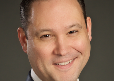 Smiling man with brown eyes and dark hair wearing a navy blue suit and black tie.