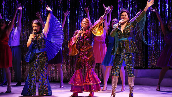Old theatre - exterior - night Photo from our 2018 production of Mamma Mia, with three performers in glittering brightly colored disco outfits, singing together.