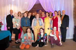 Photo from the Gala of a crowd of people lined up for a group photo. They all smile brightly in front of a sheer white drape lit with pastel pinks and blues.