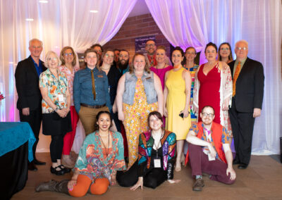 Photo from the Gala of a crowd of people lined up for a group photo. They all smile brightly in front of a sheer white drape lit with pastel pinks and blues.