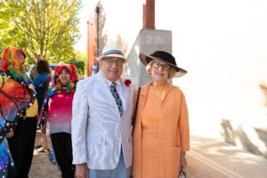 Photo of a couple at the Gala, man dressed in a light blue suit and hat and woman dressed in orange cream dress and jacket with a large black sun hat.