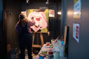 Photo from the Gala of a young woman painting an abstract and colorful painting on a roughly 24 inch by 24 inch canvas.