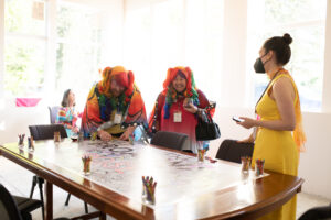 Photo from the Gala of two rainbow-dressed attendees (complete with rainbow wigs) helping color in a large banner spread across a table. A woman in a bright yellow dress watches.