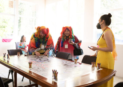 Photo from the Gala of two rainbow-dressed attendees (complete with rainbow wigs) helping color in a large banner spread across a table. A woman in a bright yellow dress watches.