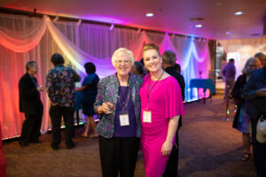 Photo from the Gala in the theatre lobby. Shows two women side by side, one dressed in bold pink and the other in dark blue. They are smiling brightly for the camera.