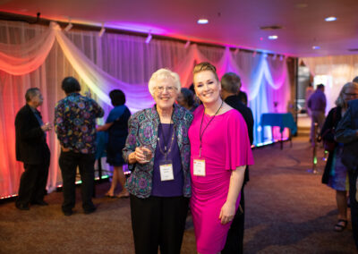 Photo from the Gala in the theatre lobby. Shows two women side by side, one dressed in bold pink and the other in dark blue. They are smiling brightly for the camera.