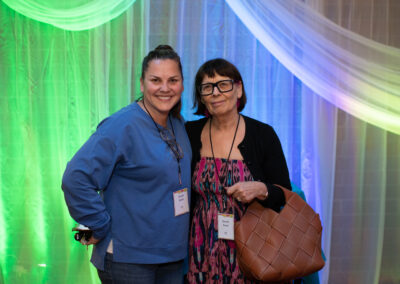 Photo from the Gala of two women, side by side, smiling brightly for the camera. They are standing in front of a white, sheer drape brightly lit with greens and blues.