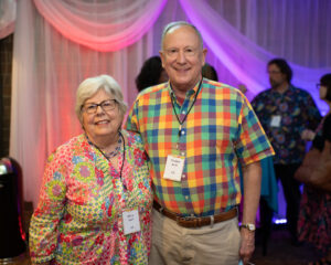 Photo from the Gala in the theatre lobby of two attendees, a man and woman, standing side by side smiling brightly for the camera. They are wearing colorful plaid and floral-printed clothing.