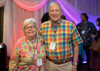 Photo from the Gala in the theatre lobby of two attendees, a man and woman, standing side by side smiling brightly for the camera. They are wearing colorful plaid and floral-printed clothing.