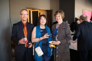 Photo from the Gala of three attendees, a man and two women, standing side by side and smiling brightly for the camera. They are in the new rehearsal hall near the double doors leading to the offices.