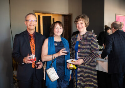 Photo from the Gala of three attendees, a man and two women, standing side by side and smiling brightly for the camera. They are in the new rehearsal hall near the double doors leading to the offices.