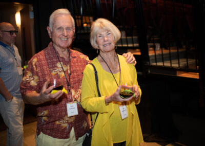 Photo from the Gala of a man and woman, side by side, his arm around her shoulder. He is wearing a deep red Hawaiian shirt and she is wearing a bright yellow shirt and sweater.