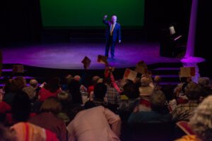 Photo from the Gala of a man on the stage wearing a black suit with an orange tie holding his right hand in the air. The crowd is holding up their programs in the air.