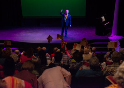 Photo from the Gala of a man on the stage wearing a black suit with an orange tie holding his right hand in the air. The crowd is holding up their programs in the air.