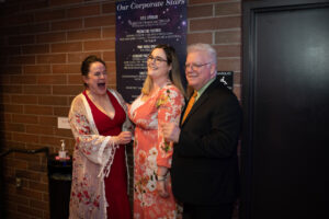 Photo from the Gala of three attendees standing in a row in front of a brown brick wall. From left to right, a woman in a red dress with a sheer floral robe on top, a woman in a peach-colored floral printed dress, and a man in a black suit and orange tie. The first woman is laughing while the other two wait for their picture to be taken.