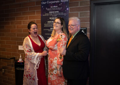 Photo from the Gala of three attendees standing in a row in front of a brown brick wall. From left to right, a woman in a red dress with a sheer floral robe on top, a woman in a peach-colored floral printed dress, and a man in a black suit and orange tie. The first woman is laughing while the other two wait for their picture to be taken.