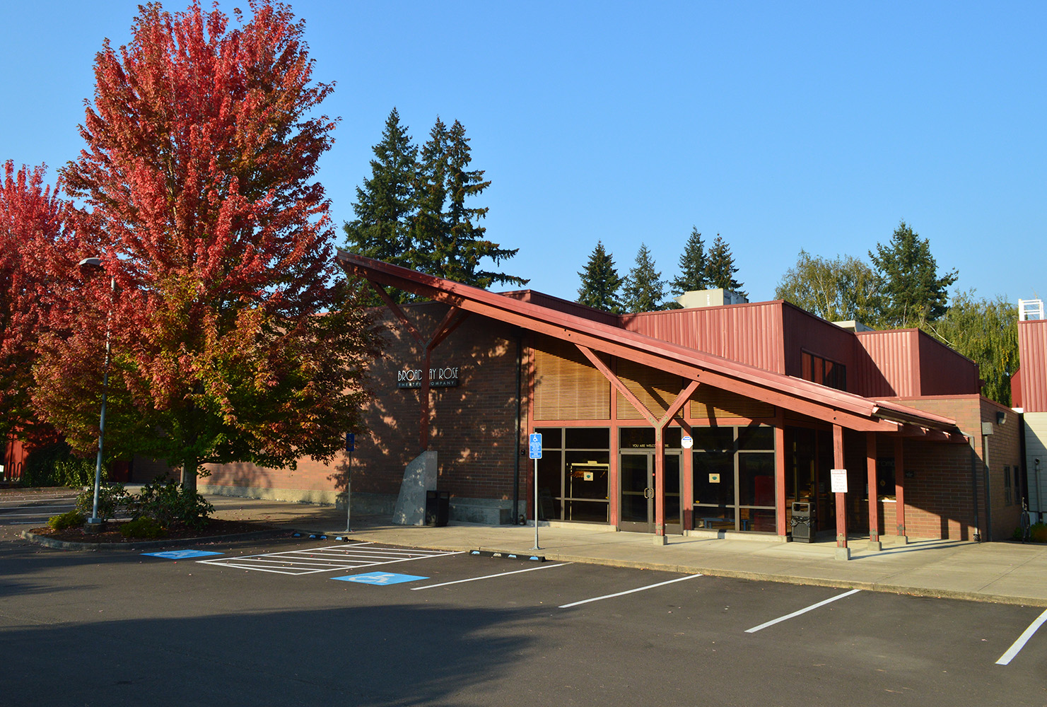 Broadway Rose New Stage Theatre, Fall 2022. Brilliant red and orange leaves cover the trees towering above the entrance to the lobby.