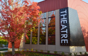 Photo of the exterior of Broadway Rose New Stage Theatre showing the large, light-up sign reading "Theatre" and the vibrant red leaves of the tree in front of the building.