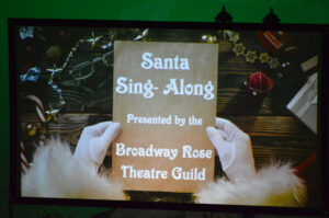 Photo of the lyrics screen showing the pre-show image: Santa holding some parchment paper that reads: Santa Sing-Along, presented by the Broadway Rose Theatre Guild.