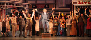 Photo from The Music Man (2014), of Harold Hill in front of a crowd of singing townsfolk.