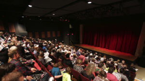 Photo of the interior of the New Stage Theater, the audience is full, the stage lights are on, lighting up the red main curtain.