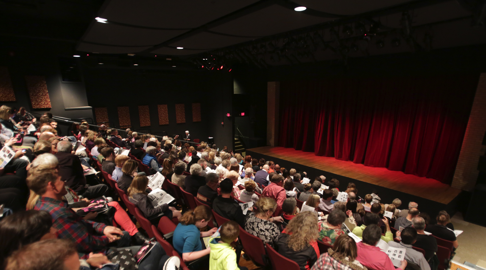 Photo of the interior of the New Stage Theater, the audience is full, the stage lights are on, lighting up the red main curtain.