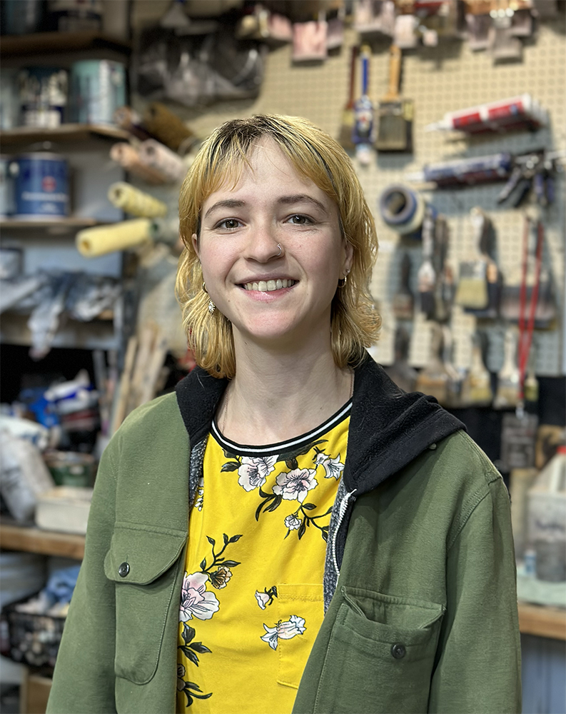 Elaine Akamian, wearing a hooded army-green jacket, standing in front of a workbench covered in painting and sculpting materials.