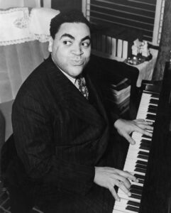 Photo of Thomas Fats Waller playing a piano and looking back over his shoulder, smiling coyly.
