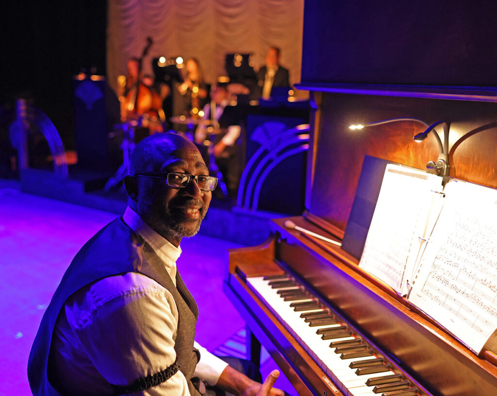 William Knowles sitting at his piano on stage left, smiling for the camera, with the rest of the band in the background.