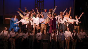 Photo from our production of Big Fish, showing the end of the song "Be the Hero". The entire cast surrounds a young Will Bloom and lifts him over their shoulders triumphantly.